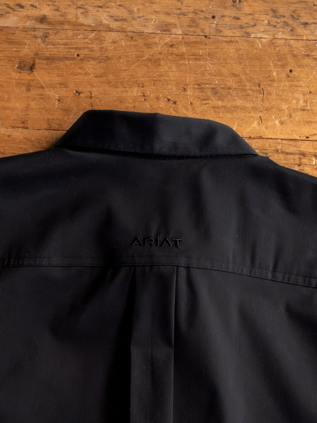 The Newnan Ariat Men's Black Button Up by Savannah Sevens western life{&}style features ARIAT embroidery on the upper back. This wrinkle resistant shirt, shown on a wooden surface, offers both style and comfort.