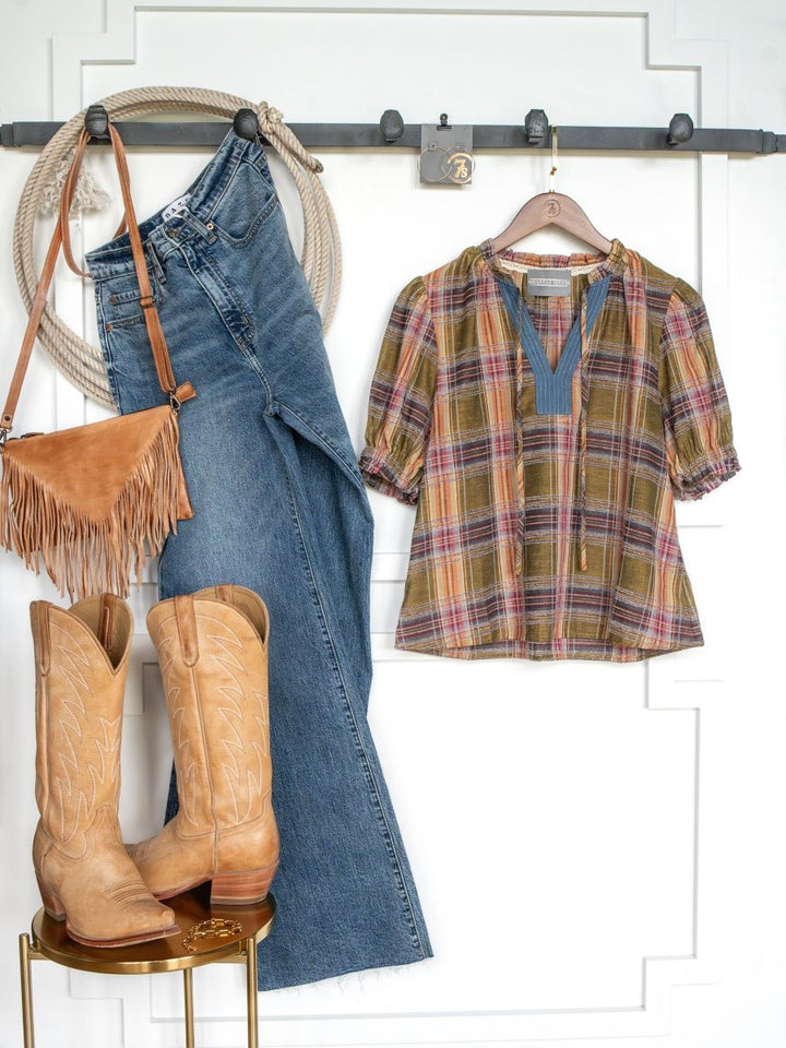 A Western outfit featuring blue jeans, the Nicoma Plaid Puff Sleeve Top by Savannah Sevens western life{&}style, a tan fringe bag, tan cowboy boots, and a coiled rope displayed on hooks and a small gold table against a white paneled wall.