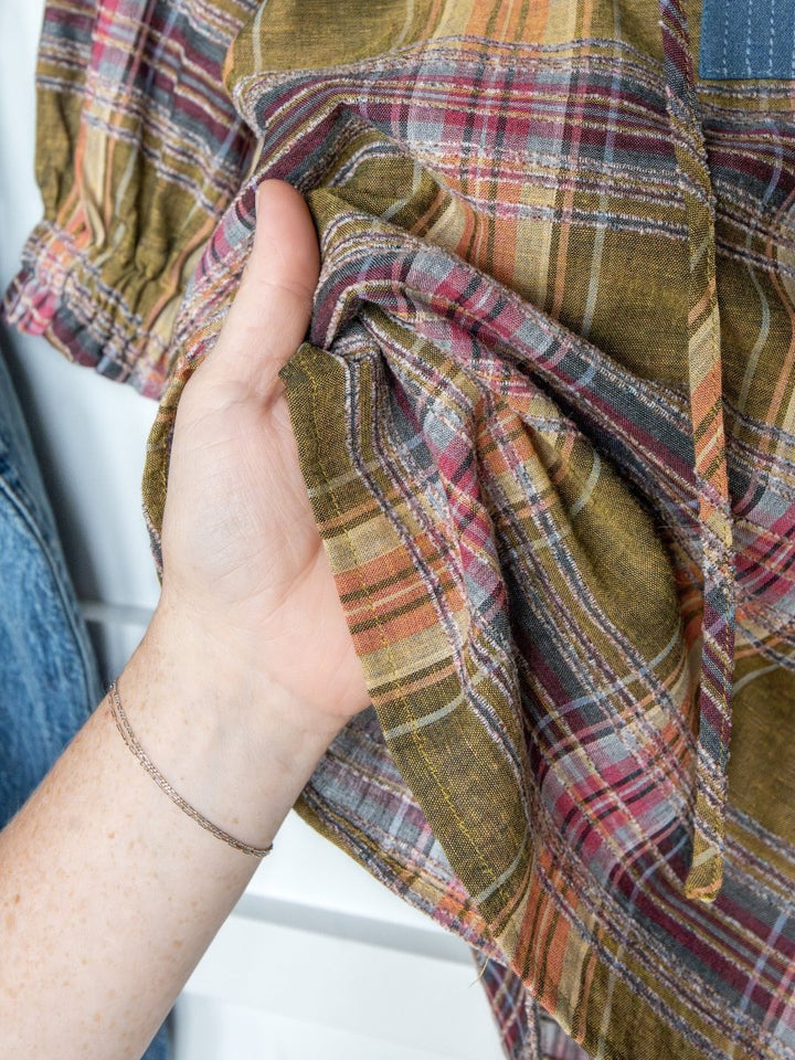 A hand with a delicate double bracelet gently gathers the Nicoma Plaid Puff Sleeve Top by Savannah Sevens western life{&}style, showcasing its soft multicolored plaid in olive green, red, yellow, and blue.