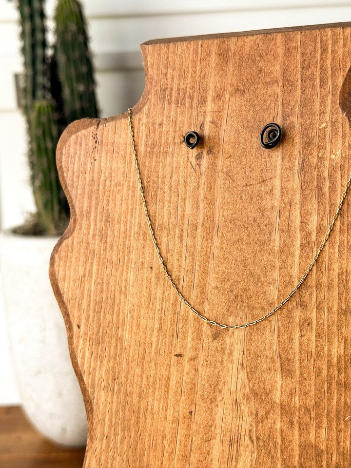 The Niota Necklace by Savannah Sevens western life{&}style is a thin, tarnish-resistant 14k gold filled chain displayed on a wooden stand, with a blurred cactus in a white pot in the background.