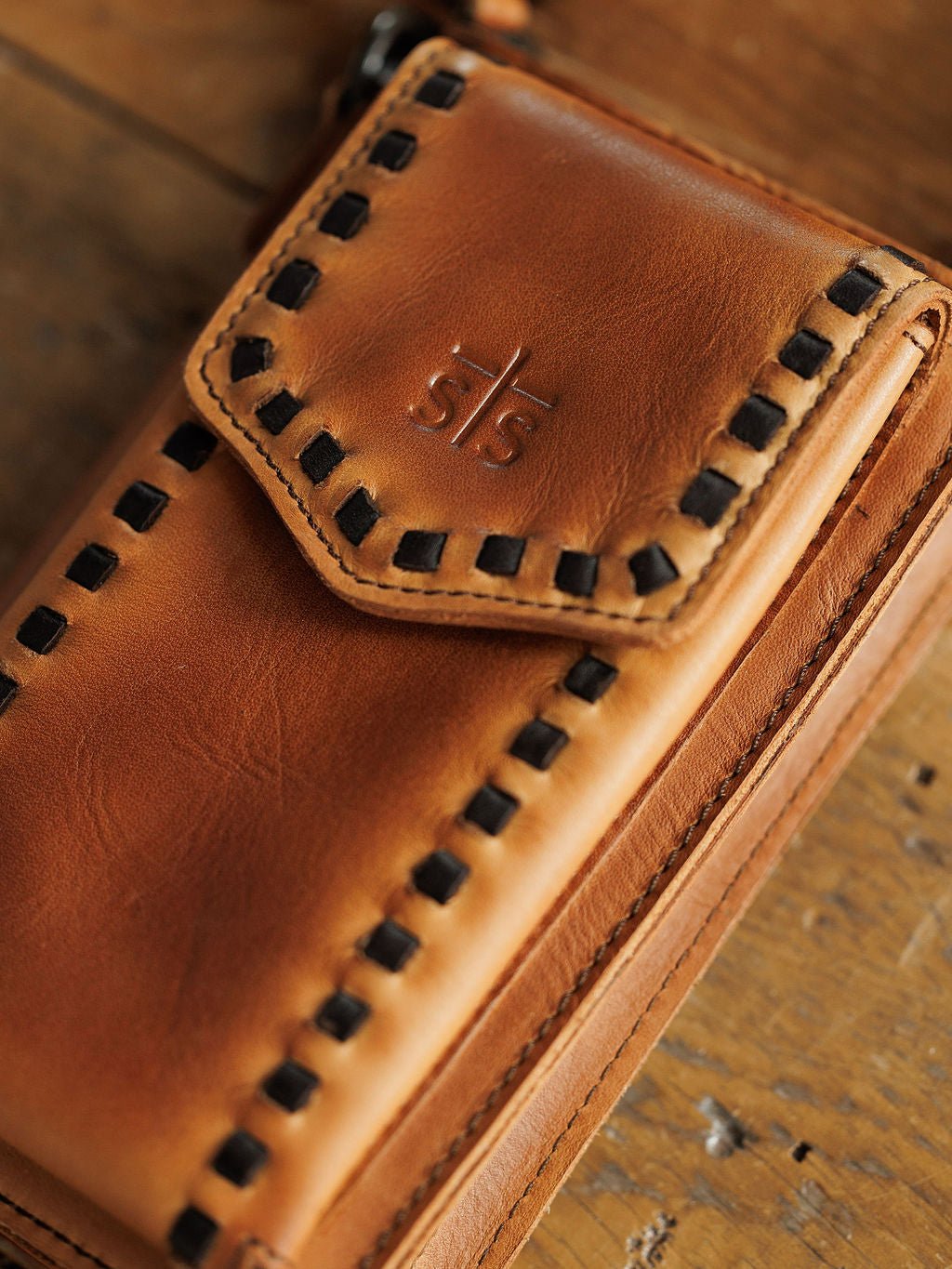Close-up of a brown Nocona Leather Crossbody Wallet by Savannah Sevens western life{&}style with black stitched edges and an embossed logo on the flap, resting on a wooden surface. By STS.