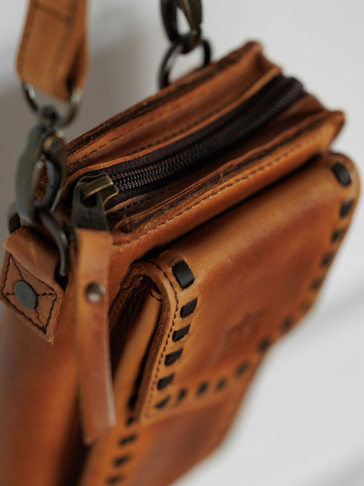 A close-up of the Savannah Sevens western life{&}style Nocona Leather Crossbody Wallet shows brown full grain leather with black stitching, a zipper closure, a metal clasp on the strap, and a smooth polished finish. By STS.