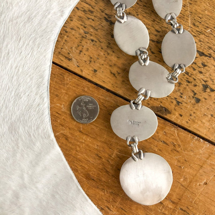 The Nogales Necklace by Savannah Sevens western life{&}style, featuring large flat oval and round sterling silver links, is displayed on wood beside a U.S. quarter with white cowhide partially visible on the left.