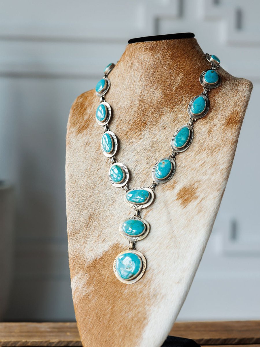 The Nogales Necklace by Savannah Sevens western life{&}style features handmade Kingman turquoise and sterling silver oval stones, displayed on a brown and white cowhide stand with a softly blurred background.