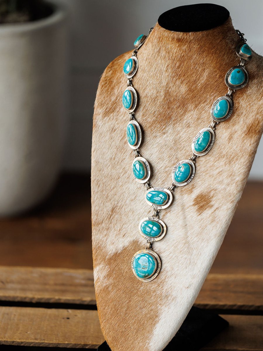 The Nogales Necklace by Savannah Sevens western life{&}style, featuring large Kingman turquoise stones, is showcased on a brown and white cowhide stand atop a wooden surface with a blurred plant pot in the background.