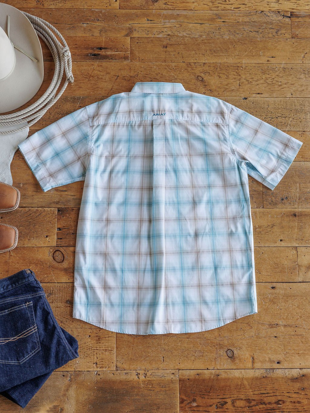 The Nolanville Ariat Men's Plaid Button Up by Savannah Sevens western life{&}style, a light blue and white short-sleeve shirt, is displayed flat on a wooden floor with a cowboy hat, brown sandals, and folded denim jeans nearby.