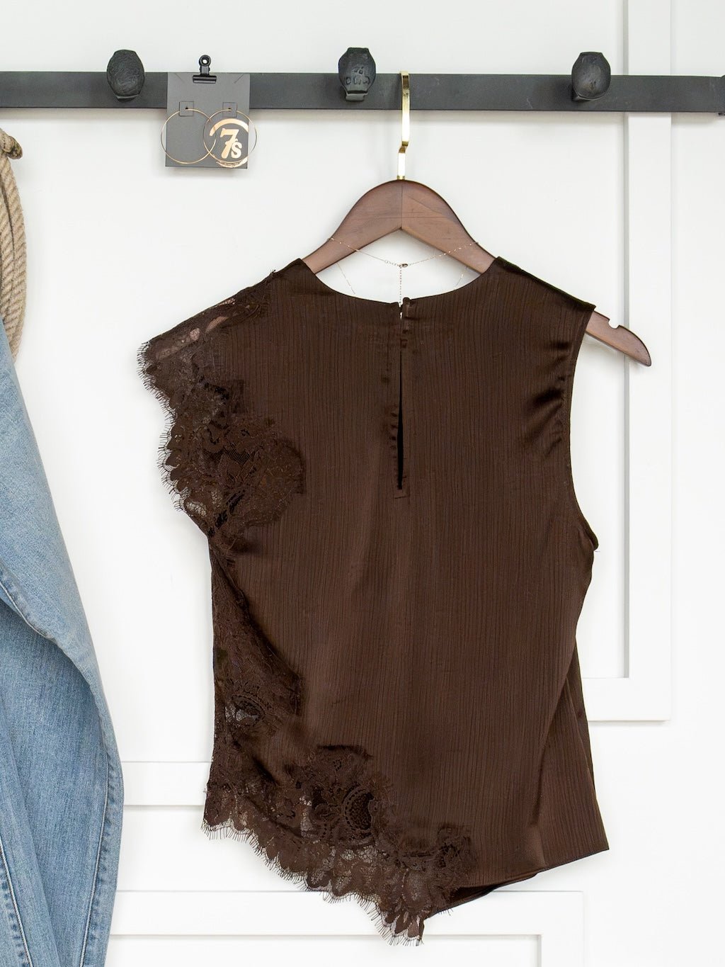 The Nora Chocolate Lace Trim Top by Savannah Sevens western life{&}style, a sleeveless piece featuring lace detail on one side, hangs on a wooden hanger against a white wall. Part of a blue denim jacket is visible on the left.