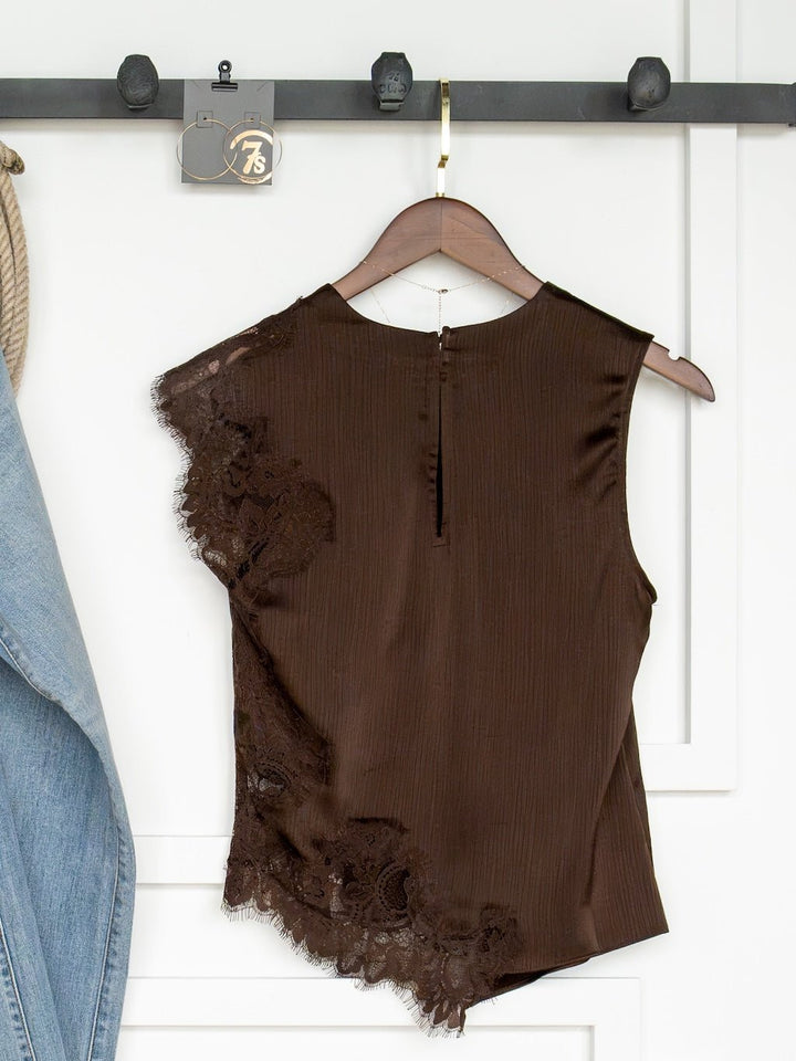 The Nora Chocolate Lace Trim Top by Savannah Sevens western life{&}style, a sleeveless piece featuring lace detail on one side, hangs on a wooden hanger against a white wall. Part of a blue denim jacket is visible on the left.