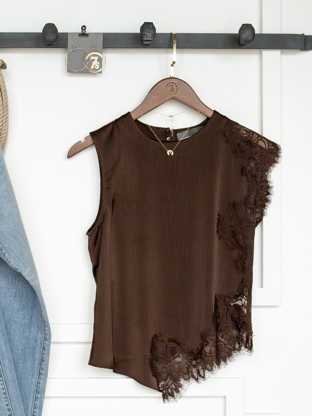 The Nora Chocolate Lace Trim Top by Savannah Sevens western life{&}style—a sleeveless dark chocolate top featuring an asymmetrical hem and delicate lace trim on one side—hangs on a wooden hanger from a black rack. A denim jacket is partially visible to the left, with a necklace draped from the same hook.