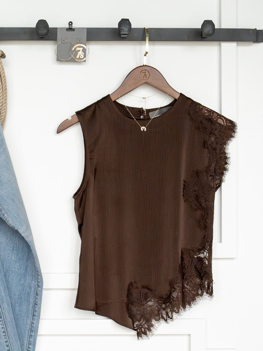 The Nora Chocolate Lace Trim Top by Savannah Sevens western life{&}style—a sleeveless dark chocolate top featuring an asymmetrical hem and delicate lace trim on one side—hangs on a wooden hanger from a black rack. A denim jacket is partially visible to the left, with a necklace draped from the same hook.