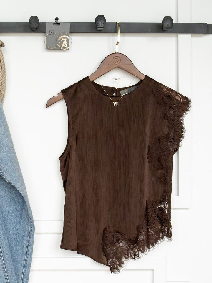 The Nora Chocolate Lace Trim Top by Savannah Sevens western life{&}style—a sleeveless dark chocolate top featuring an asymmetrical hem and delicate lace trim on one side—hangs on a wooden hanger from a black rack. A denim jacket is partially visible to the left, with a necklace draped from the same hook.
