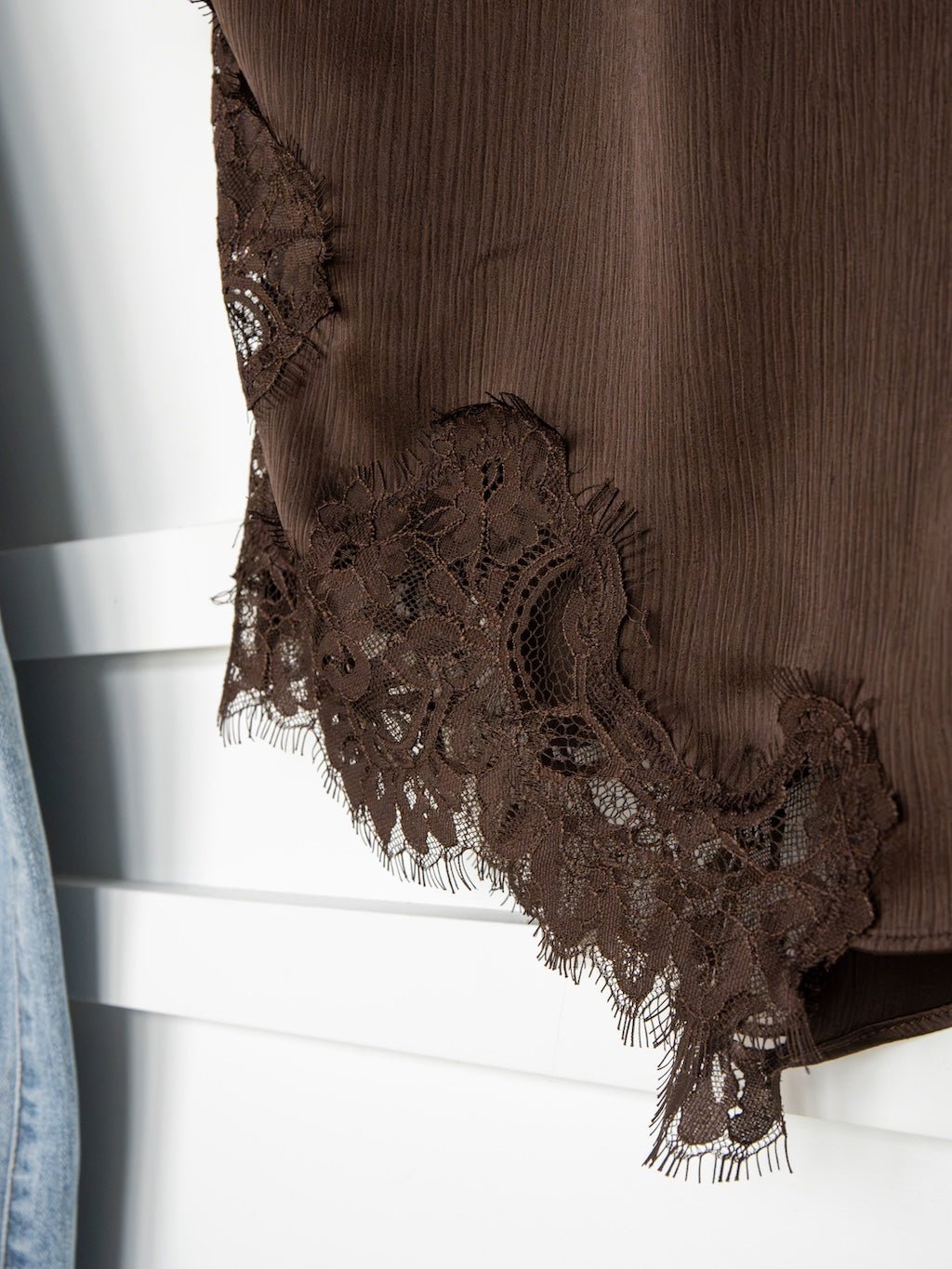 Close-up of the Nora Chocolate Lace Trim Top by Savannah Sevens western life{&}style, featuring delicate lace trim along the hem, displayed alongside a pair of light blue jeans against a white background.