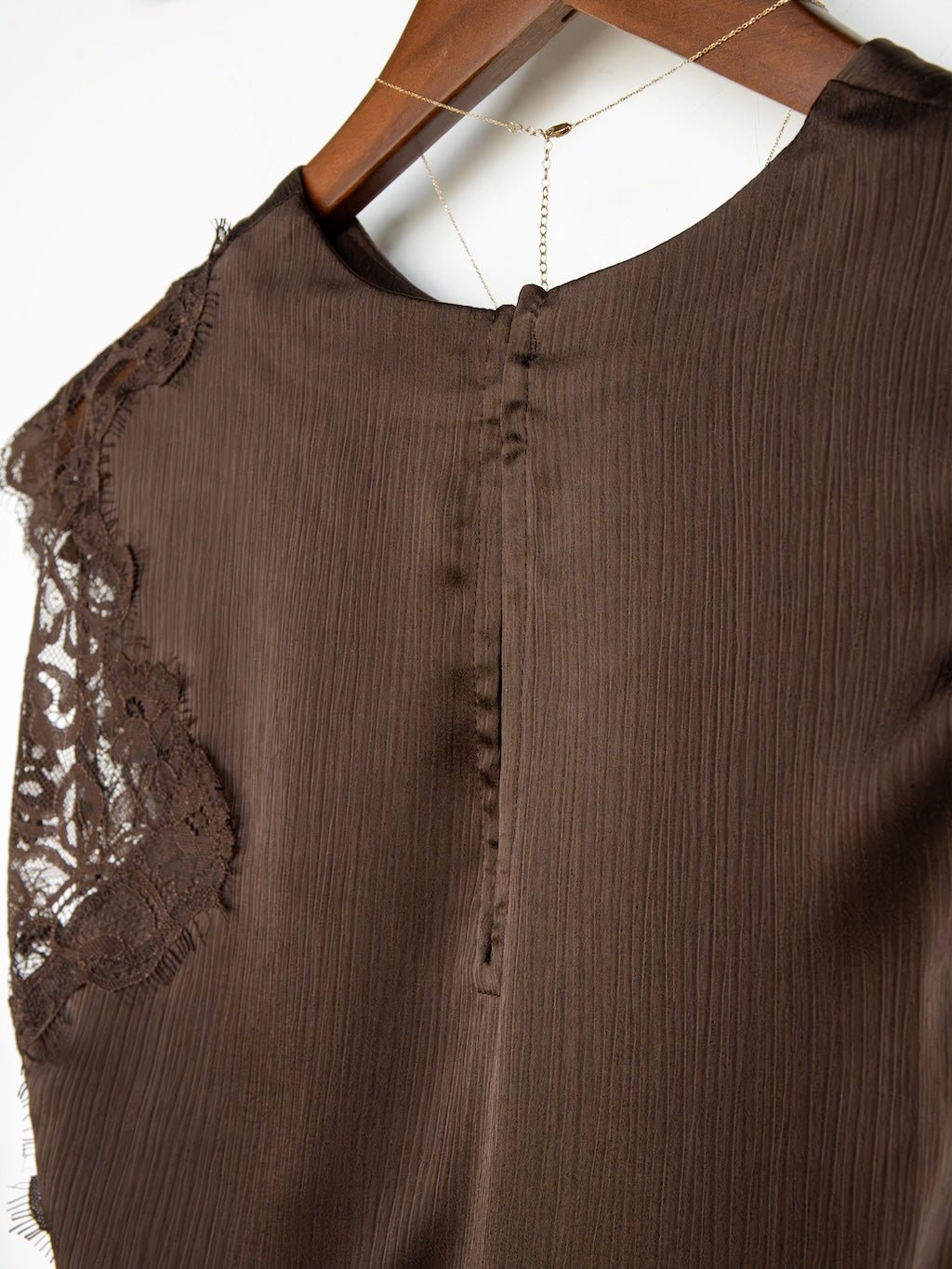 Close-up of the back of the Savannah Sevens western life{&}style Nora Chocolate Lace Trim Top, a dark chocolate sleeveless piece featuring delicate lace detail on one shoulder, subtle textured fabric, and a hidden zipper closure, displayed on a wooden hanger.