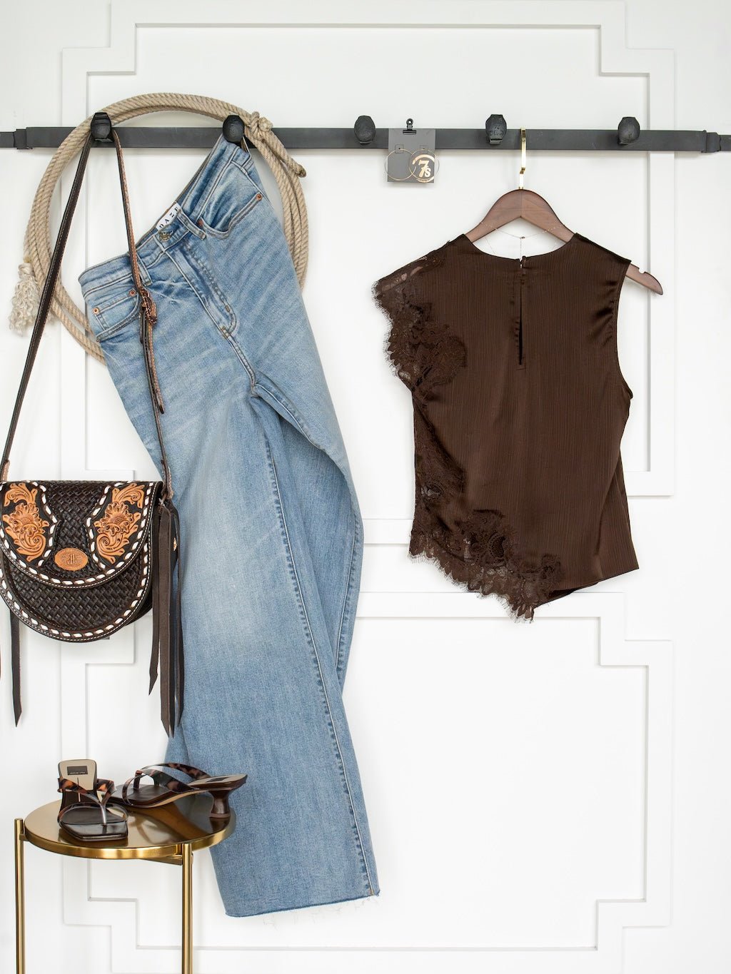 The Nora Chocolate Lace Trim Top by Savannah Sevens western life{&}style is styled with light blue jeans, a black and gold patterned handbag, black sandals, and sunglasses, all displayed neatly against a white wall.