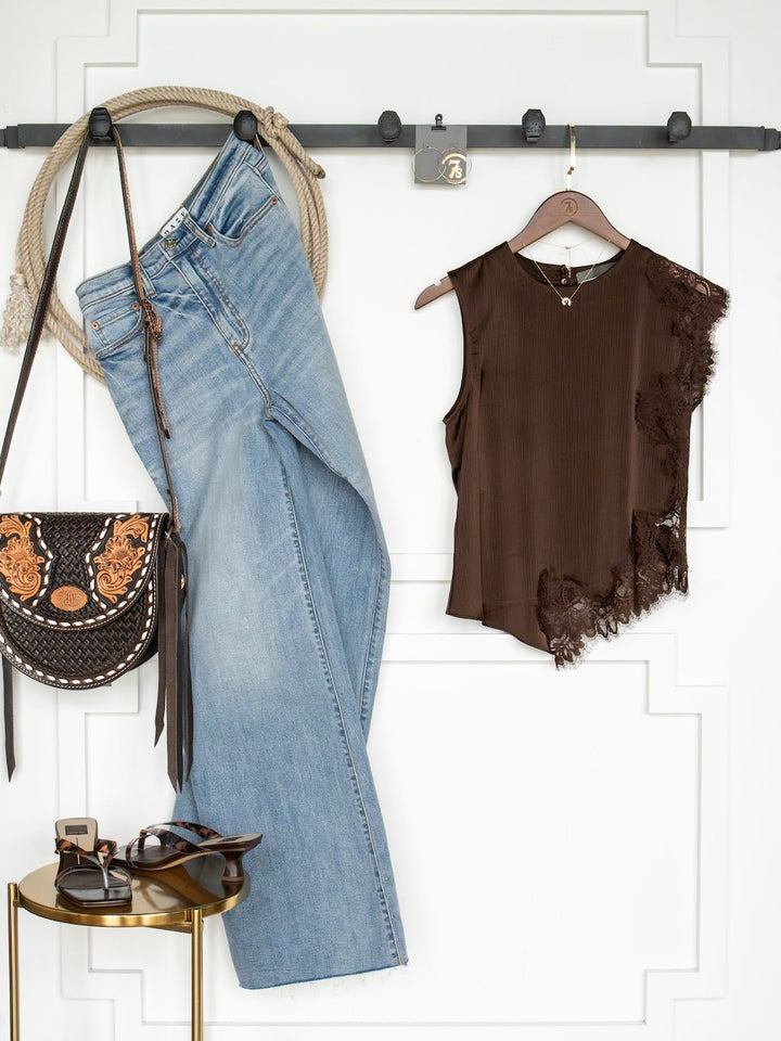 The Nora Chocolate Lace Trim Top by Savannah Sevens western life{&}style is styled with a pair of blue jeans, a black and brown purse, brown sandals, and a necklace, all displayed against a white wall with decorative molding.