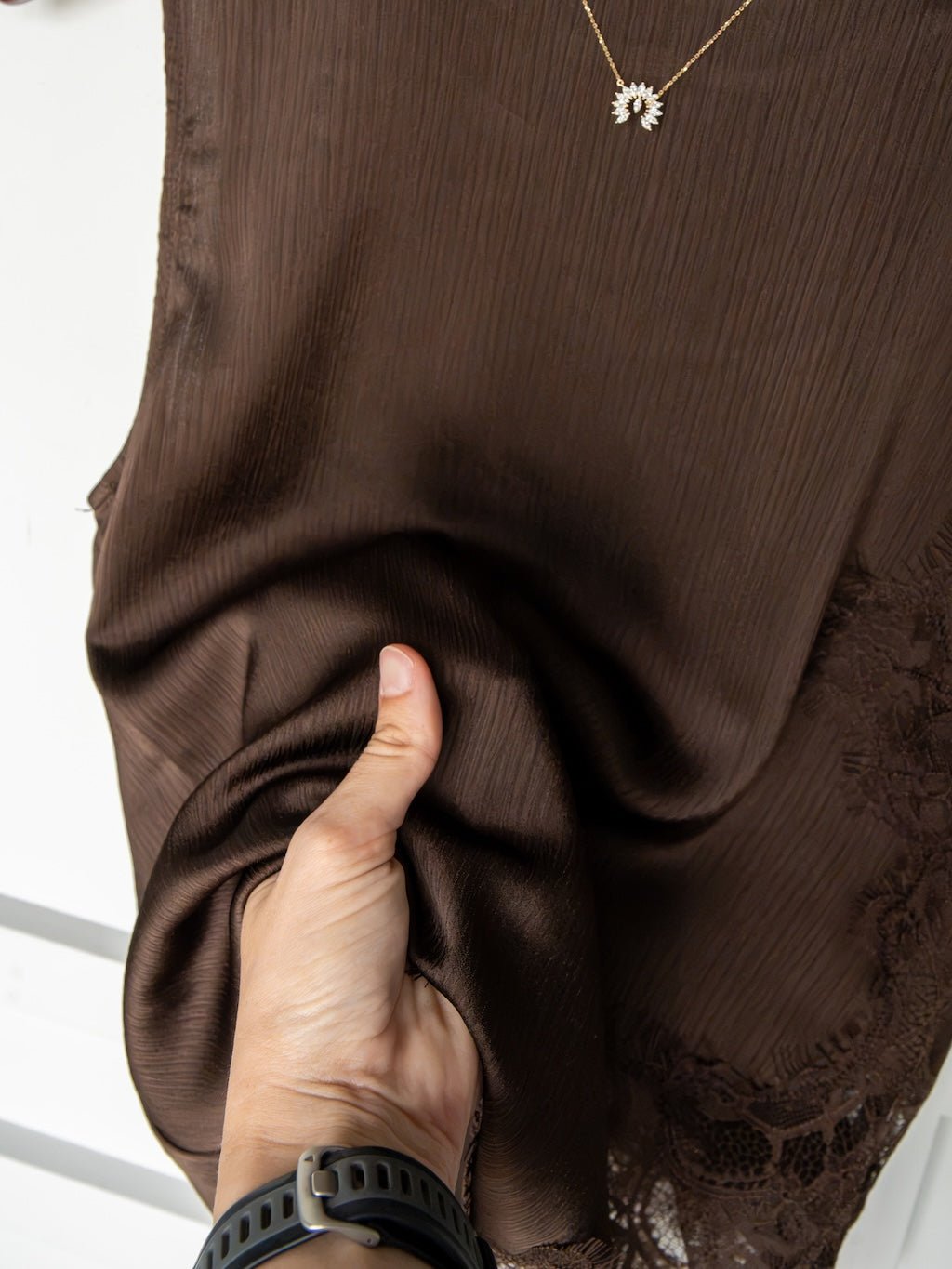 A hand holding and softly scrunching the fabric of the Nora Chocolate Lace Trim Top by Savannah Sevens western life{&}style, highlighting its delicate lace trim; the person is accessorized with a black wristband and a gold sun-shaped pendant necklace.