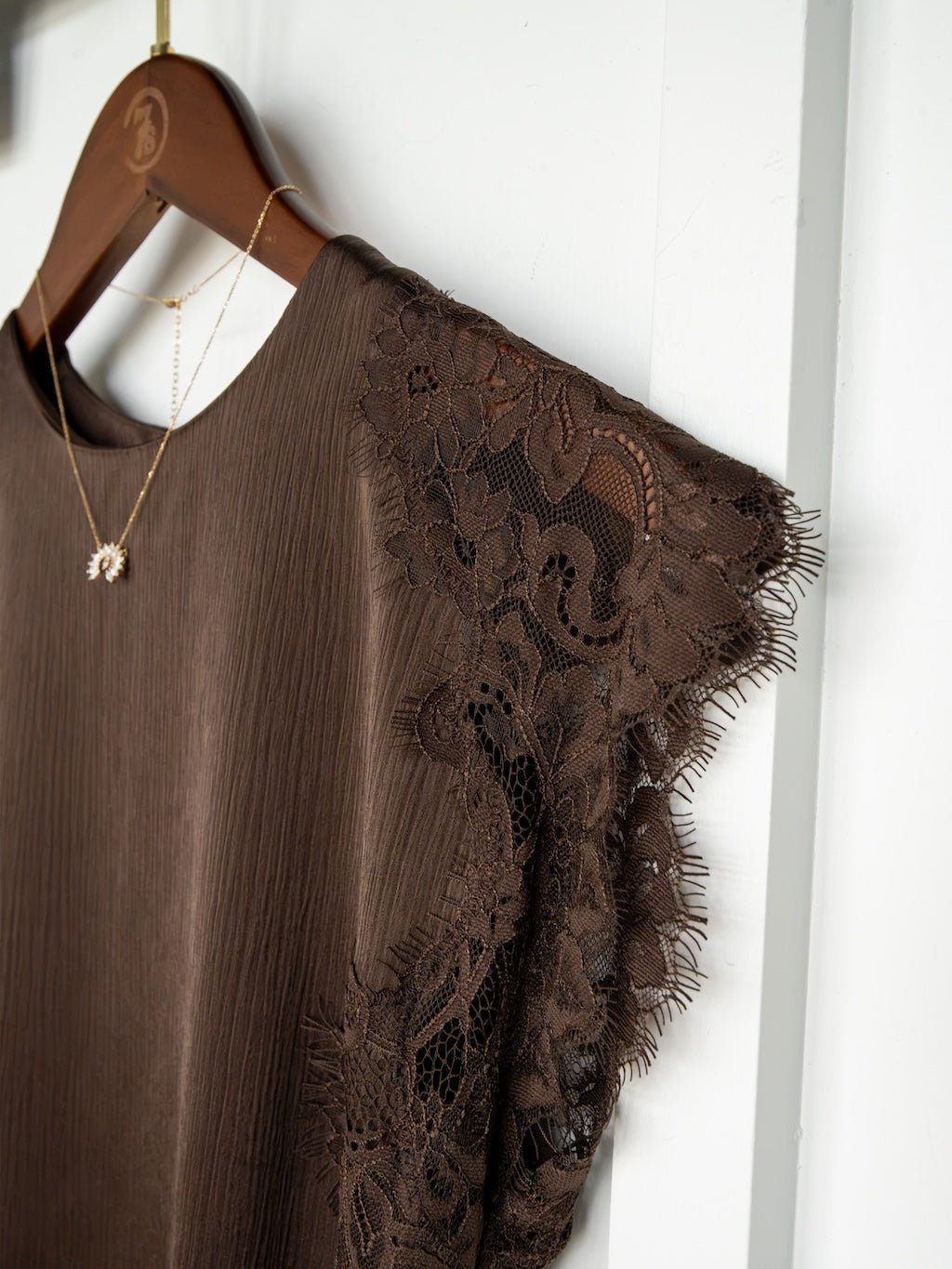 A Nora Chocolate Lace Trim Top by Savannah Sevens western life{&}style hangs on a wooden hanger, showcasing delicate lace detailing at the sleeves. It's paired with a gold necklace featuring a small pendant, creating an elegant ensemble reminiscent of satin and lace.