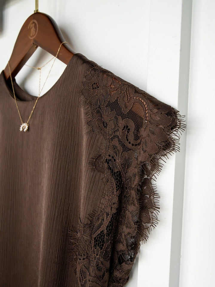 A Nora Chocolate Lace Trim Top by Savannah Sevens western life{&}style hangs on a wooden hanger, showcasing delicate lace detailing at the sleeves. It's paired with a gold necklace featuring a small pendant, creating an elegant ensemble reminiscent of satin and lace.