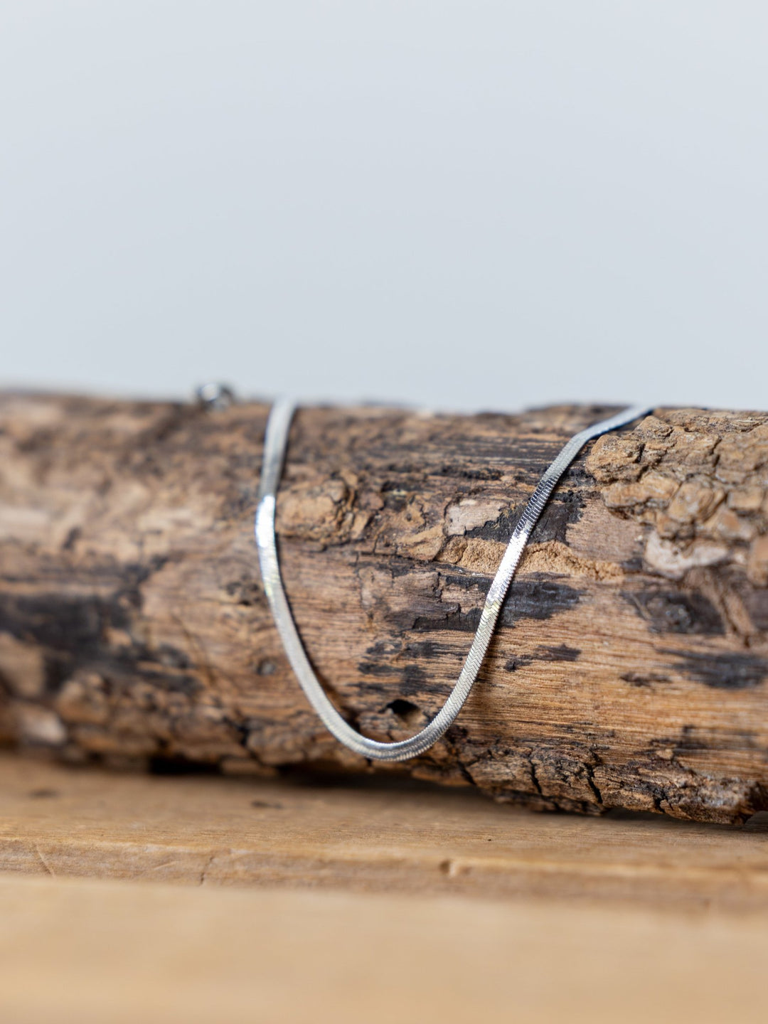 A thin silver chain necklace rests on textured, weathered wood with a neutral background, beautifully complementing the Norvell Silver Herringbone Bracelet by Savannah Sevens western life{&}style.
