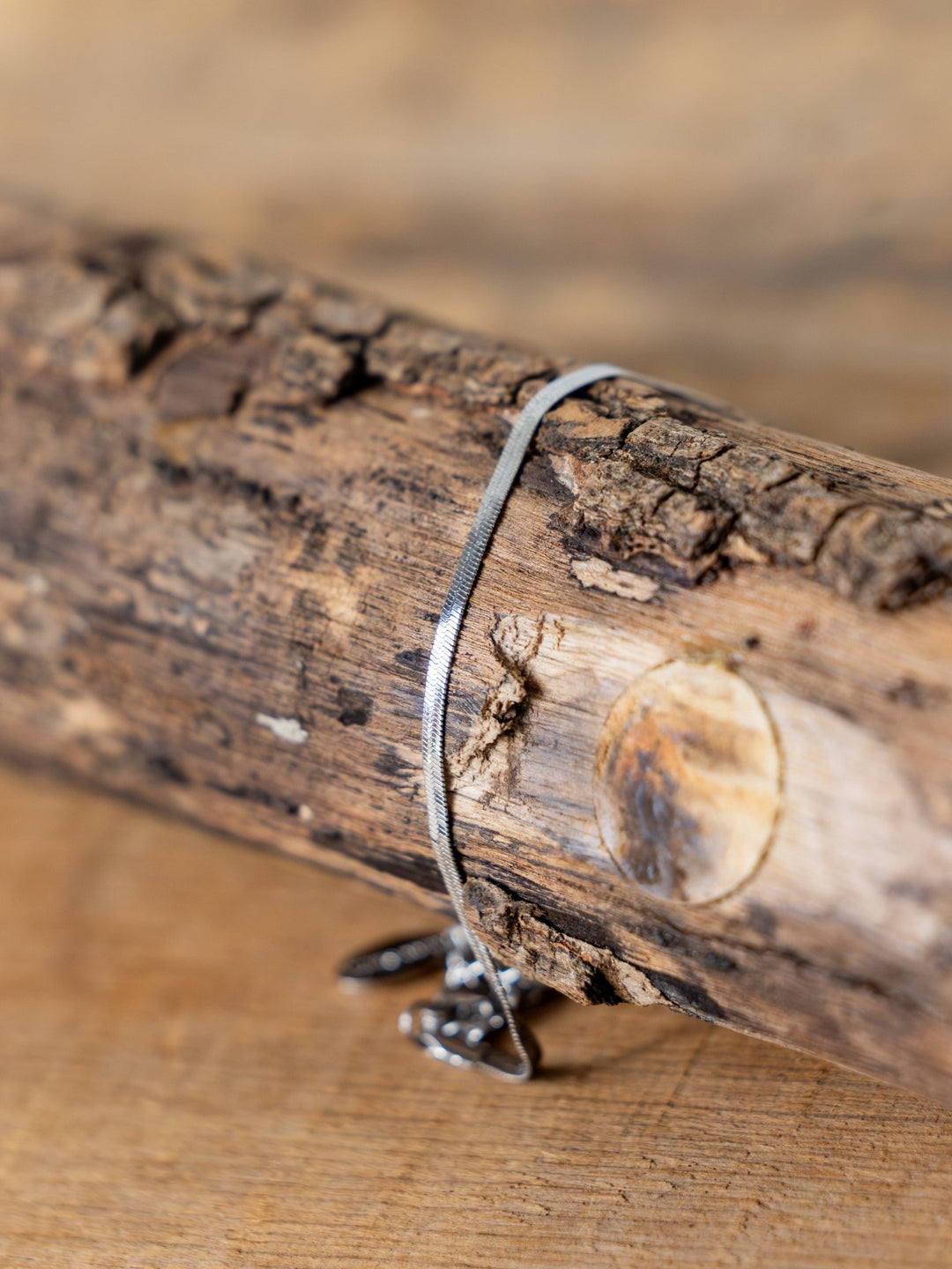 A slim silver necklace with a pendant rests on rustic wood, its grain in focus. Pair it with the Norvell Silver Herringbone Bracelet by Savannah Sevens western life{&}style for a refined, coordinated look.