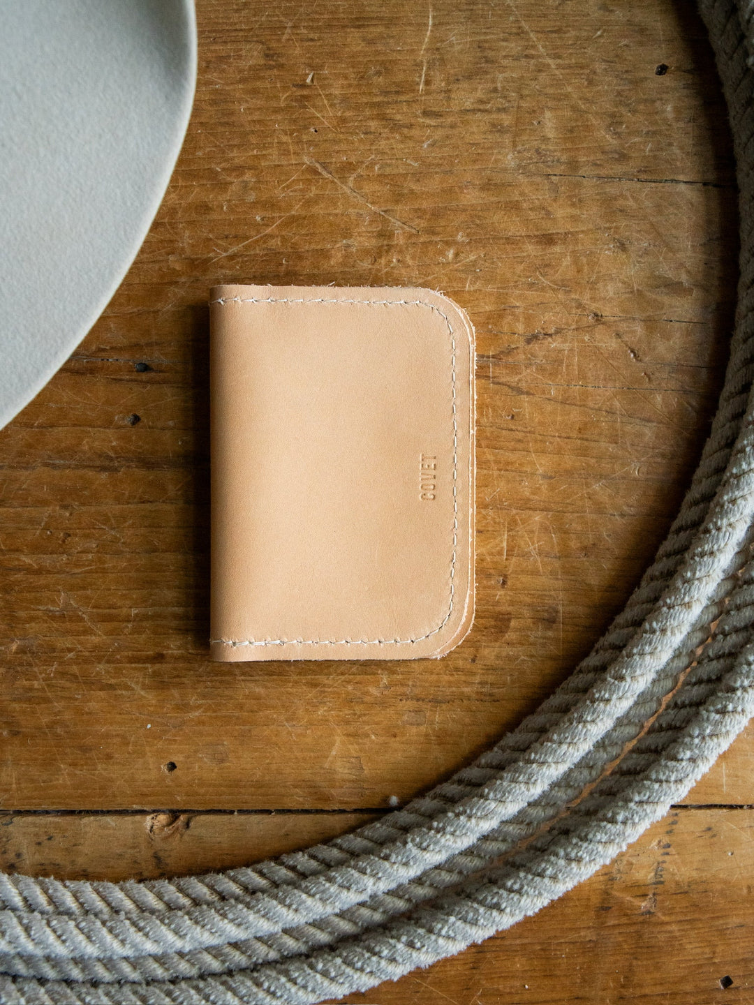 The Novell Leather Wallet by Savannah Sevens western life{&}style, crafted from full-grain leather and made in California, rests on a wooden surface. Framed by a gray rope and the edge of a light gray object in the top left corner, its tan color brings warmth to the scene.