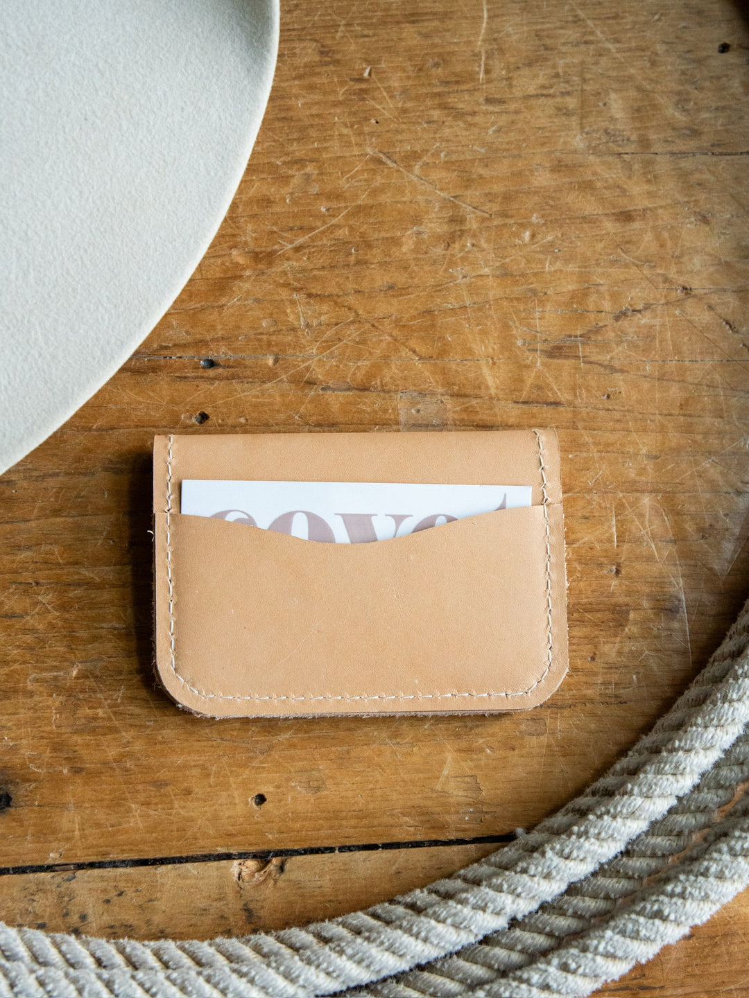 The Novell Leather Wallet by Savannah Sevens western life{&}style is a tan full-grain leather cardholder featuring visible stitching, seen here holding a partially visible card on a wooden surface beside a coiled rope and a white object in the corner. Crafted with meticulous attention to detail, this wallet emphasizes quality and durability.