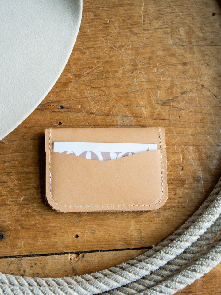 The Novell Leather Wallet by Savannah Sevens western life{&}style is a tan full-grain leather cardholder featuring visible stitching, seen here holding a partially visible card on a wooden surface beside a coiled rope and a white object in the corner. Crafted with meticulous attention to detail, this wallet emphasizes quality and durability.