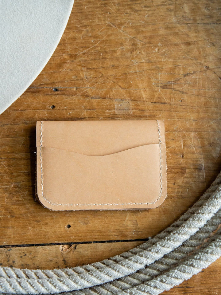 The Novell Leather Wallet by Savannah Sevens western life{&}style, crafted from tan leather in California, rests on a wooden surface beside a coiled rope and a light-colored curved object.