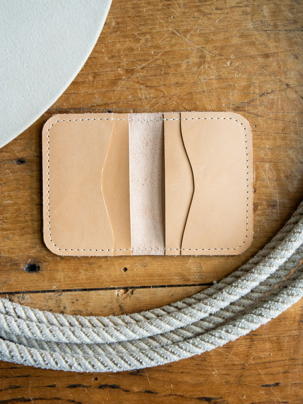 The Novell Leather Wallet by Savannah Sevens western life{&}style, crafted from tan full-grain leather with visible stitching, rests on a wooden surface beside a coiled white rope and a round white object.