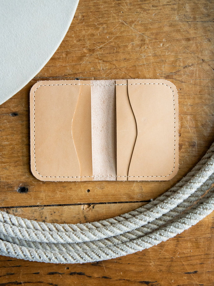 The Novell Leather Wallet by Savannah Sevens western life{&}style, crafted from tan full-grain leather with visible stitching, rests on a wooden surface beside a coiled white rope and a round white object.