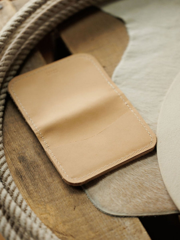 The Novell Wallet by Savannah Sevens western life{&}style is a tan full-grain leather card wallet with visible stitching, shown on cowhide and wood with a coiled rope in the background, showcasing expert craftsmanship.