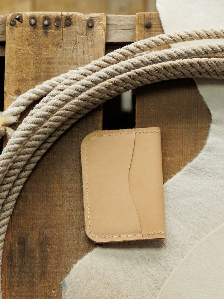 The Novell Wallet by Savannah Sevens western life{&}style, crafted from tan full-grain leather with visible stitching, is displayed on a wooden surface near a coiled rope and a white and brown cowhide.