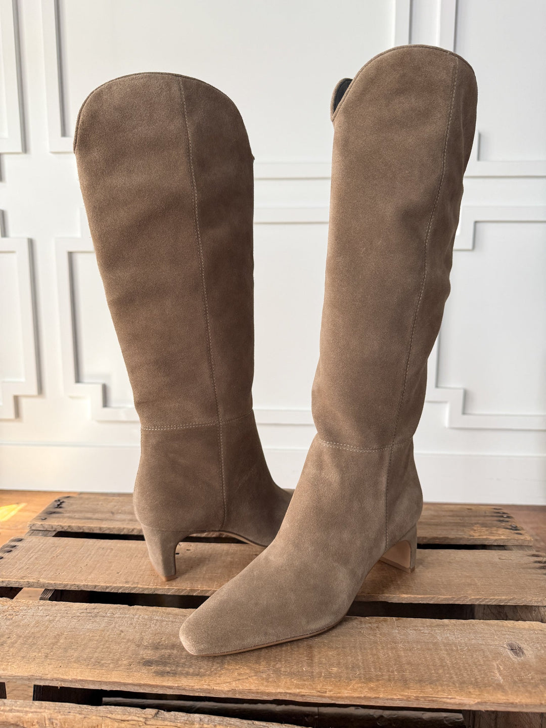 The Nyssa Taupe Suede Boot by Savannah Sevens western life{&}style—a knee-high design with pointed toes, cushioned insoles, and low heels in a distinctive shape—is displayed on a wooden pallet set against a white paneled wall.