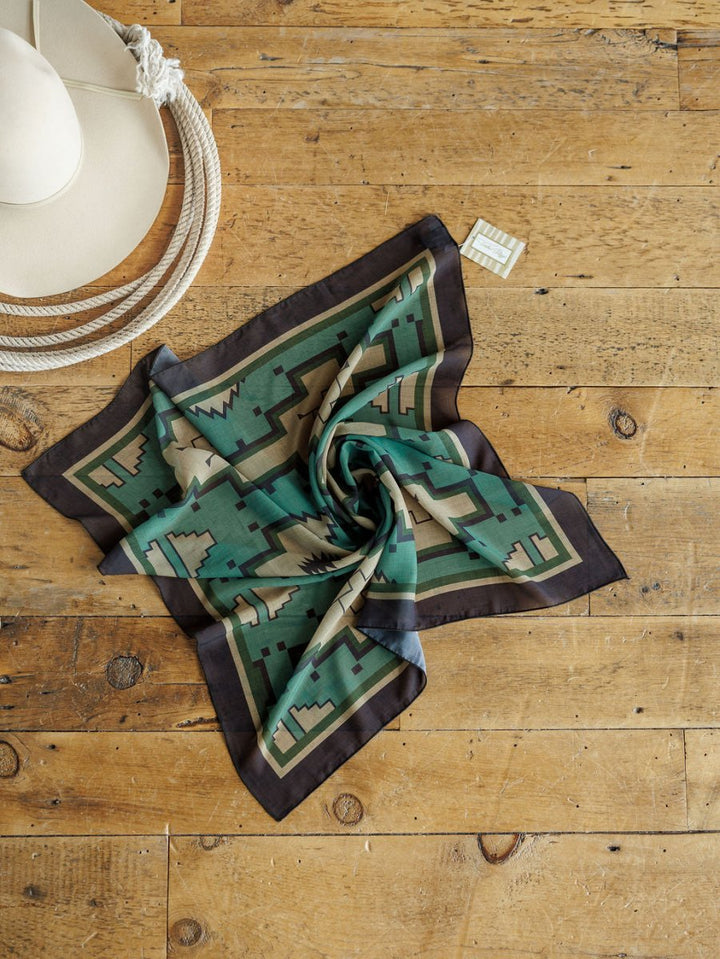 A patterned green and black bandana is neatly arranged on a rustic wooden surface beside a cream cowboy hat and the Savannah Sevens western life{&}style Oacoma Neck Scarf.
