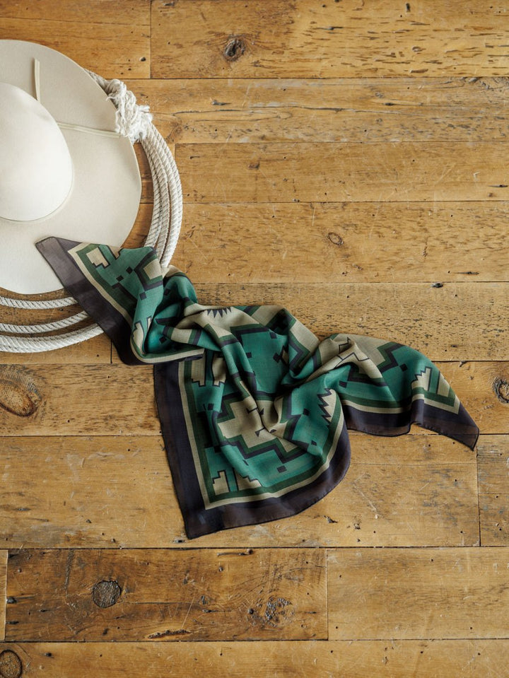 A cream-colored cowboy hat with a coiled rope and a Savannah Sevens western life{&}style Oacoma Neck Scarf rests on a rustic wooden floor.