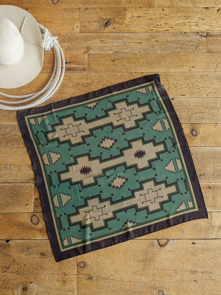 The Oacoma Neck Scarf by Savannah Sevens western life{&}style, featuring a green and beige geometric satin knit design with a black border, is displayed on a wooden floor beside a white cowboy hat.