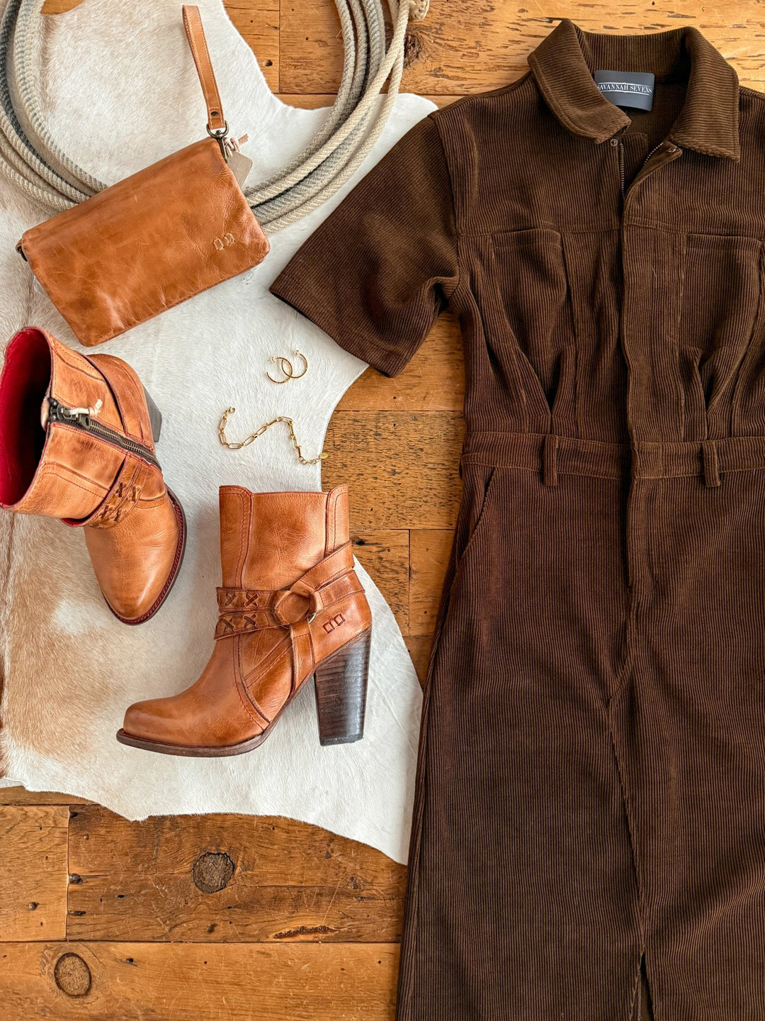 The Oak Ridge Corduroy Dress by Savannah Sevens western life{&}style is paired with tan leather heeled boots, a coordinating crossbody bag, and gold jewelry on a cowhide rug atop a rustic wooden floor.