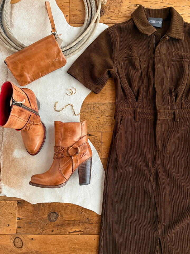 The Oak Ridge Corduroy Dress by Savannah Sevens western life{&}style is paired with tan leather heeled boots, a coordinating crossbody bag, and gold jewelry on a cowhide rug atop a rustic wooden floor.