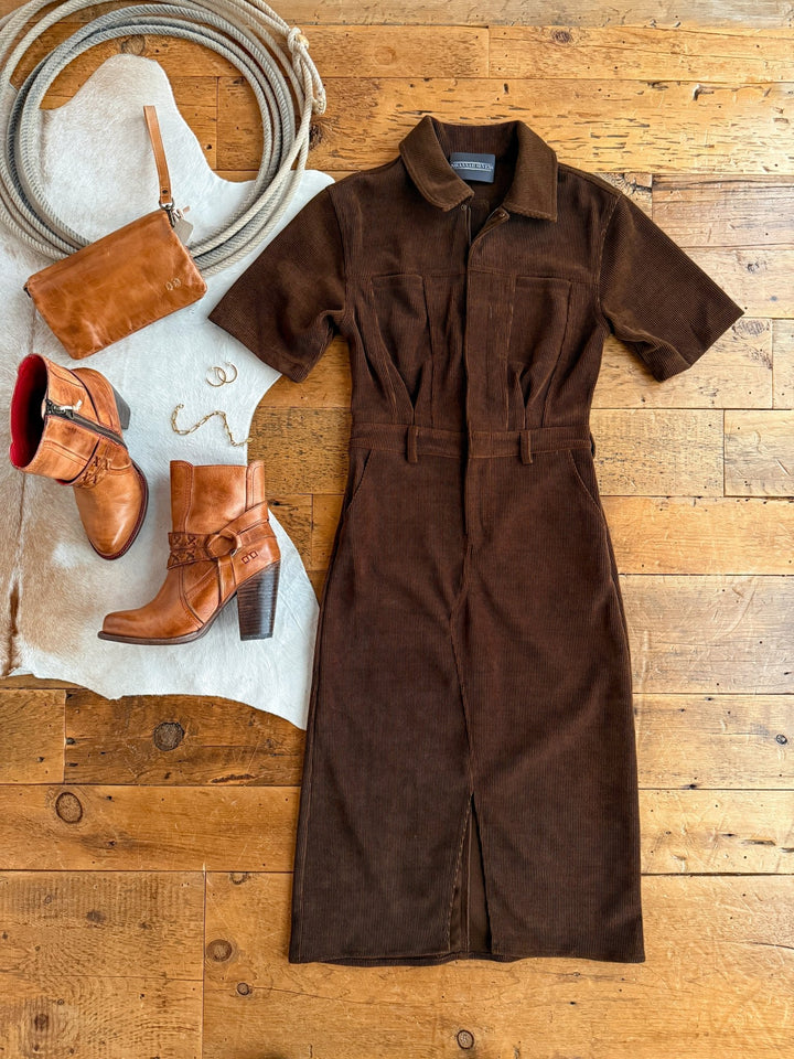 The Oak Ridge Corduroy Dress by Savannah Sevens western life{&}style is styled with tan heeled boots, a brown clutch, gold hoop earrings, and a ring on a white cowhide rug atop a wooden floor.