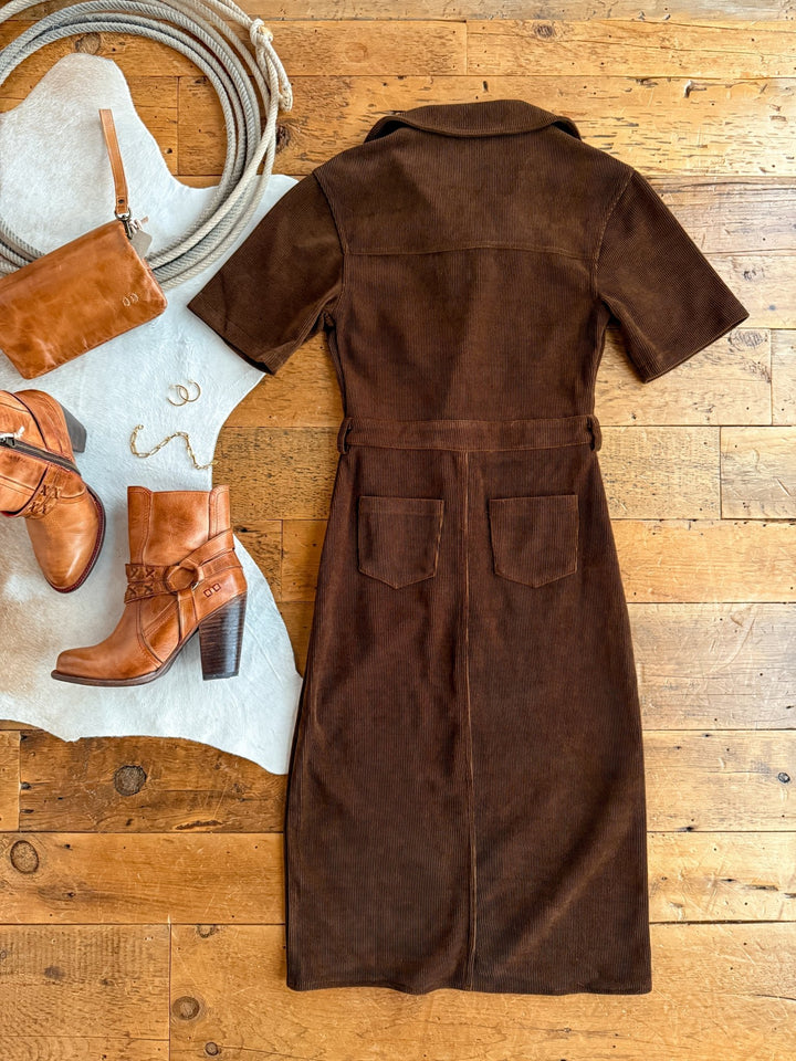 The Oak Ridge Corduroy Dress by Savannah Sevens western life{&}style, a brown short-sleeve midi, is styled on a wooden floor with brown heeled boots, a leather purse, a cowhide rug, a gold necklace, and hoop earrings.