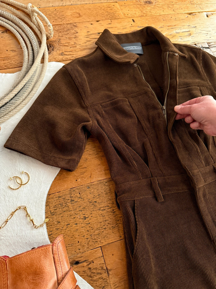 The Oak Ridge Corduroy Dress by Savannah Sevens western life{&}style is displayed on a wooden floor beside a white cowhide rug, gold jewelry, and a brown leather bag while a hand holds its zipper.