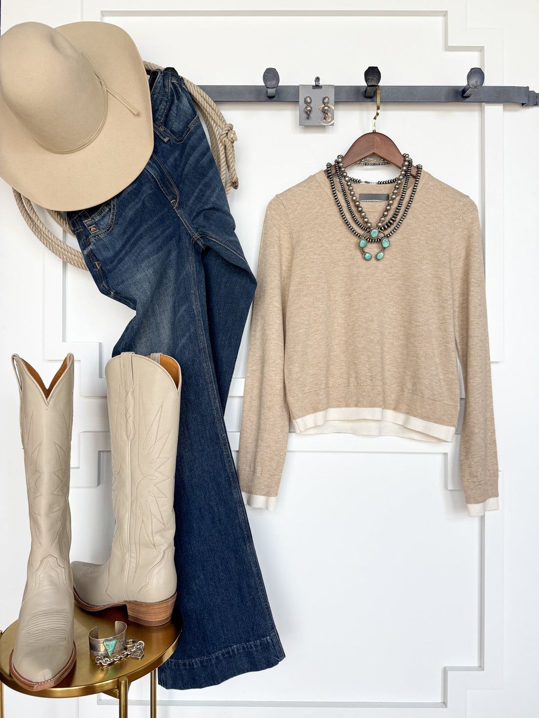 The Oakdale Oatmeal Knit Top by Savannah Sevens western life{&}style is styled on a hanger with blue jeans and a young contemporary tan cowboy hat, accented with layered necklaces. Cream cowboy boots and turquoise jewelry sit atop a gold table, all set against a white paneled wall for a modern Western-inspired look.