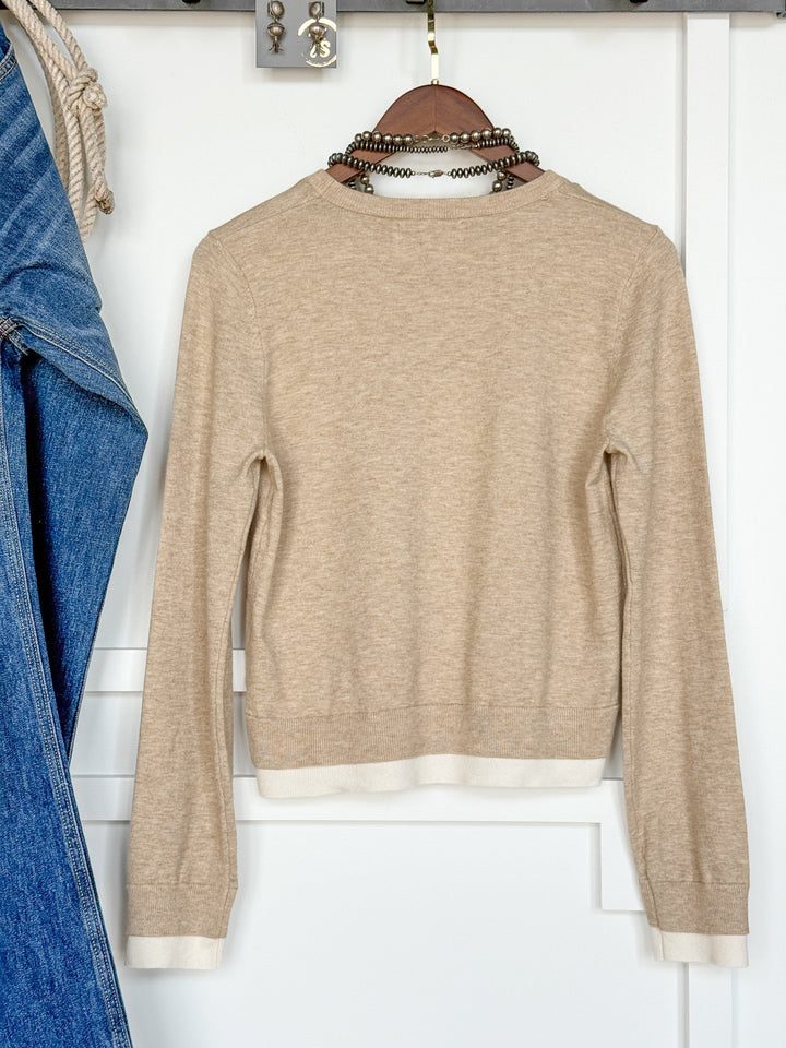 The Oakdale Oatmeal Knit Top by Savannah Sevens western life{&}style, featuring long sleeves and white trim, hangs on a wooden hanger against a white wall. A blue denim jacket is partially visible on the left side of the image.