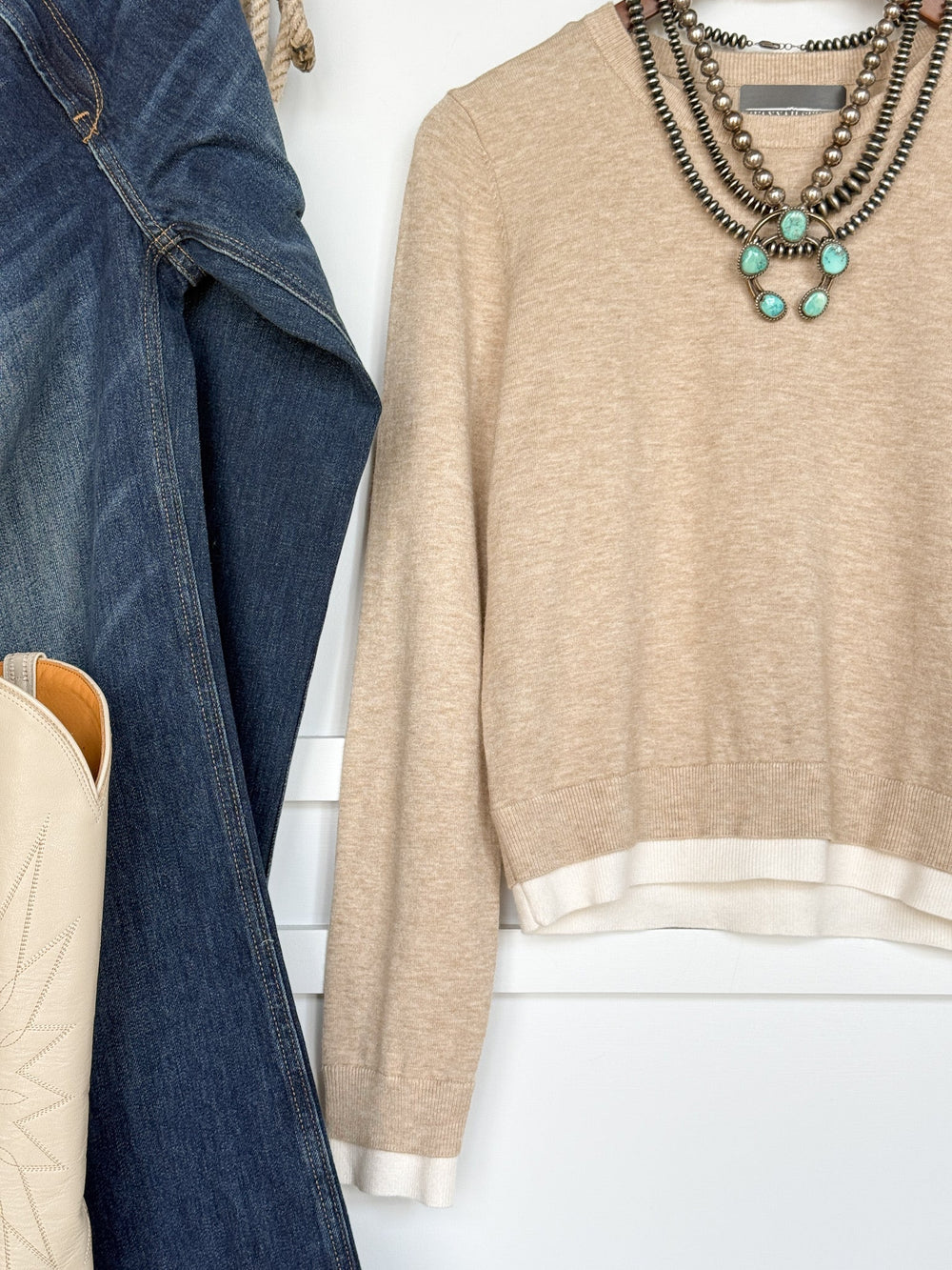 The Oakdale Oatmeal Knit Top by Savannah Sevens western life{&}style, a young contemporary beige tri-blend sweater with white trim, is styled with layered silver and turquoise necklaces, blue jeans, a denim jacket, and a cream cowboy boot hanging on a white wall.