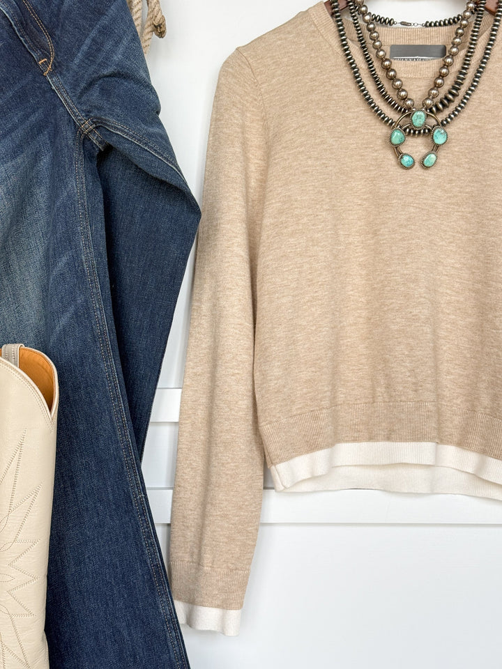 The Oakdale Oatmeal Knit Top by Savannah Sevens western life{&}style, a young contemporary beige tri-blend sweater with white trim, is styled with layered silver and turquoise necklaces, blue jeans, a denim jacket, and a cream cowboy boot hanging on a white wall.
