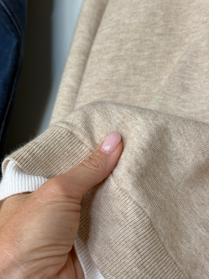 A hand with light pink polished nails is pinching the soft beige fabric of the Oakdale Oatmeal Knit Top by Savannah Sevens western life{&}style, highlighting its tri-blend knit and ribbed white cuff, set against a neutral background.