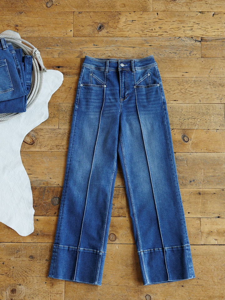 The Oakley High Rise Slim Wide Leg Jean by Savannah Sevens western life{&}style, featuring prominent front seams and large cuffs, is laid flat on a wooden floor beside a folded pair of jeans and a white cowhide rug.
