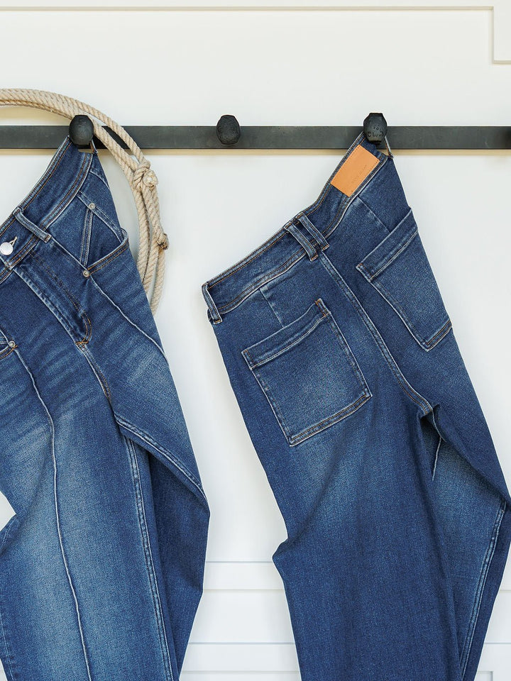 Two pairs of blue Savannah Sevens western life{&}style The Oakley High Rise Slim Wide Leg Jeans hang on black wall hooks; one pair is draped with a coiled rope, while the other neatly displays its back pockets and leather patch.