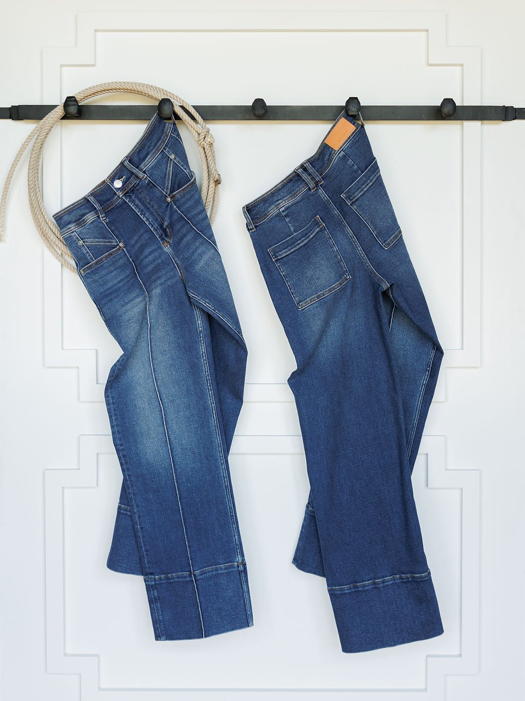 Two pairs of blue jeans, including Savannah Sevens western life{&}style's The Oakley High Rise Slim Wide Leg Jean, hang from a black rod on a white wall with trim—one pair is secured by rope and the other by a wooden hanger.