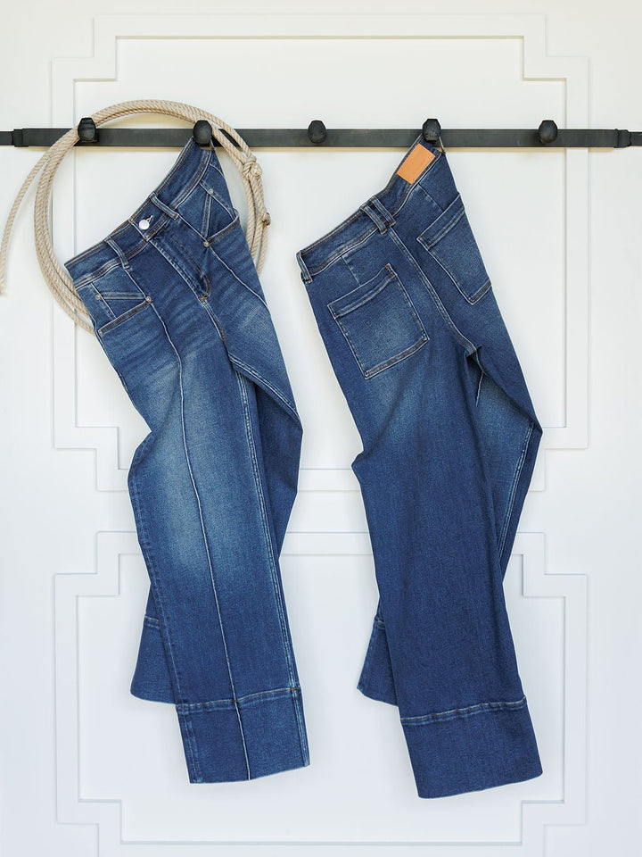Two pairs of blue jeans, including Savannah Sevens western life{&}style's The Oakley High Rise Slim Wide Leg Jean, hang from a black rod on a white wall with trim—one pair is secured by rope and the other by a wooden hanger.